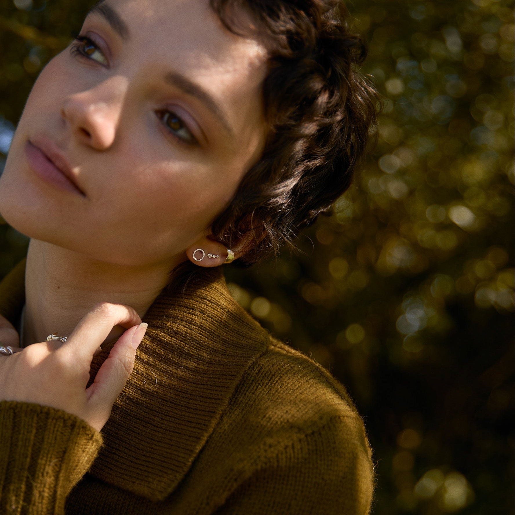 Woman wearing a brown sweater and silver circle studs with trees in the background