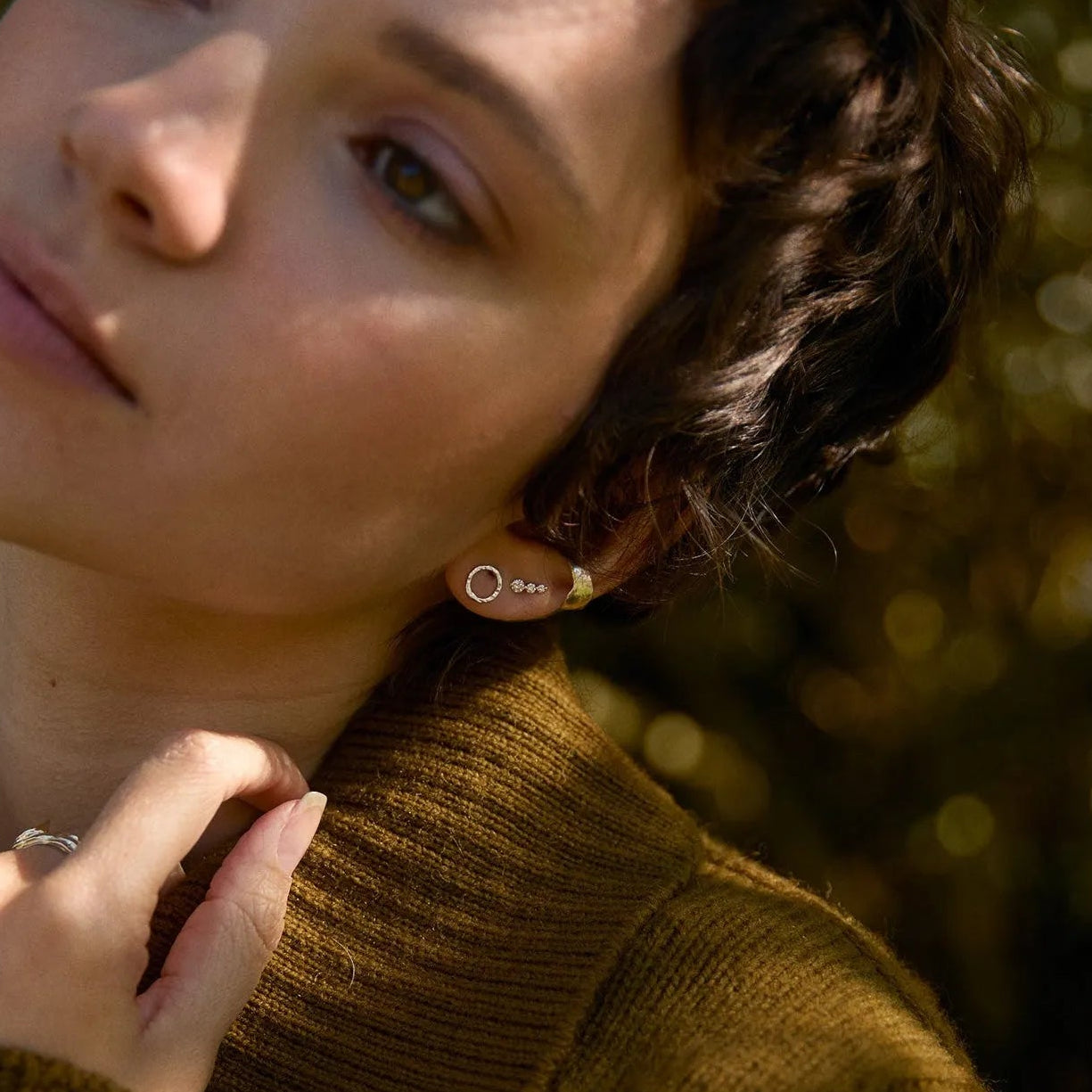 Woman wearing a brown sweater and silver circle studs with trees in the background