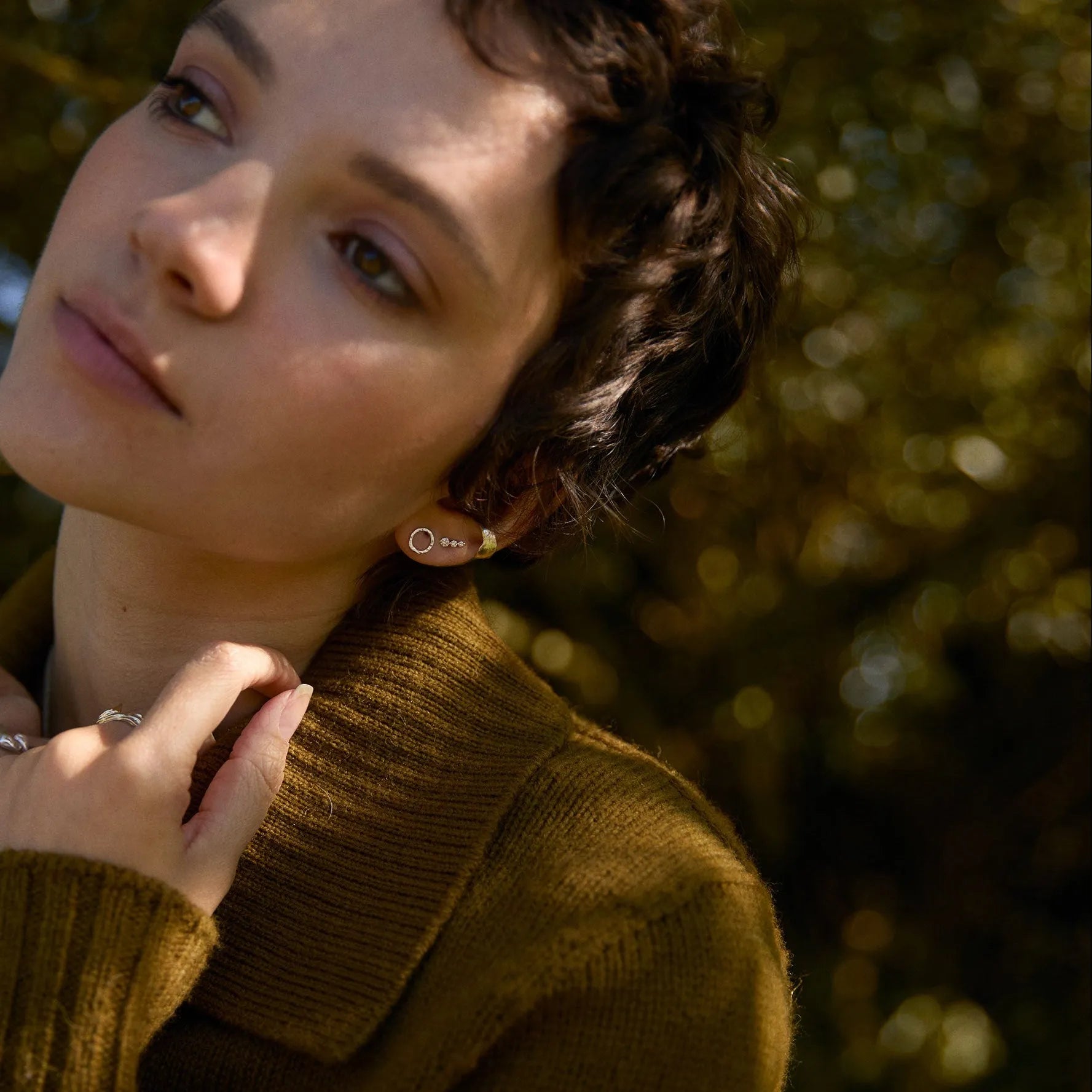 Woman wearing a brown sweater and silver circle studs with trees in the background