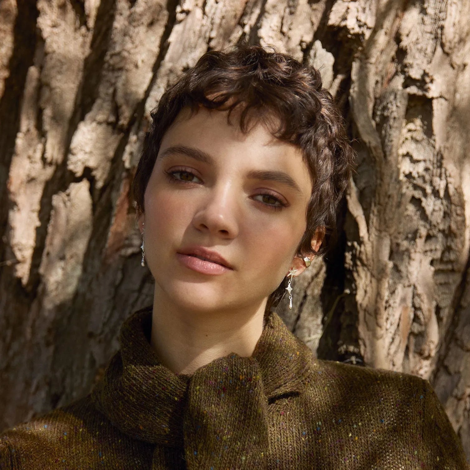 Person wearing a brown coat and star dangle earrings standing against a tree