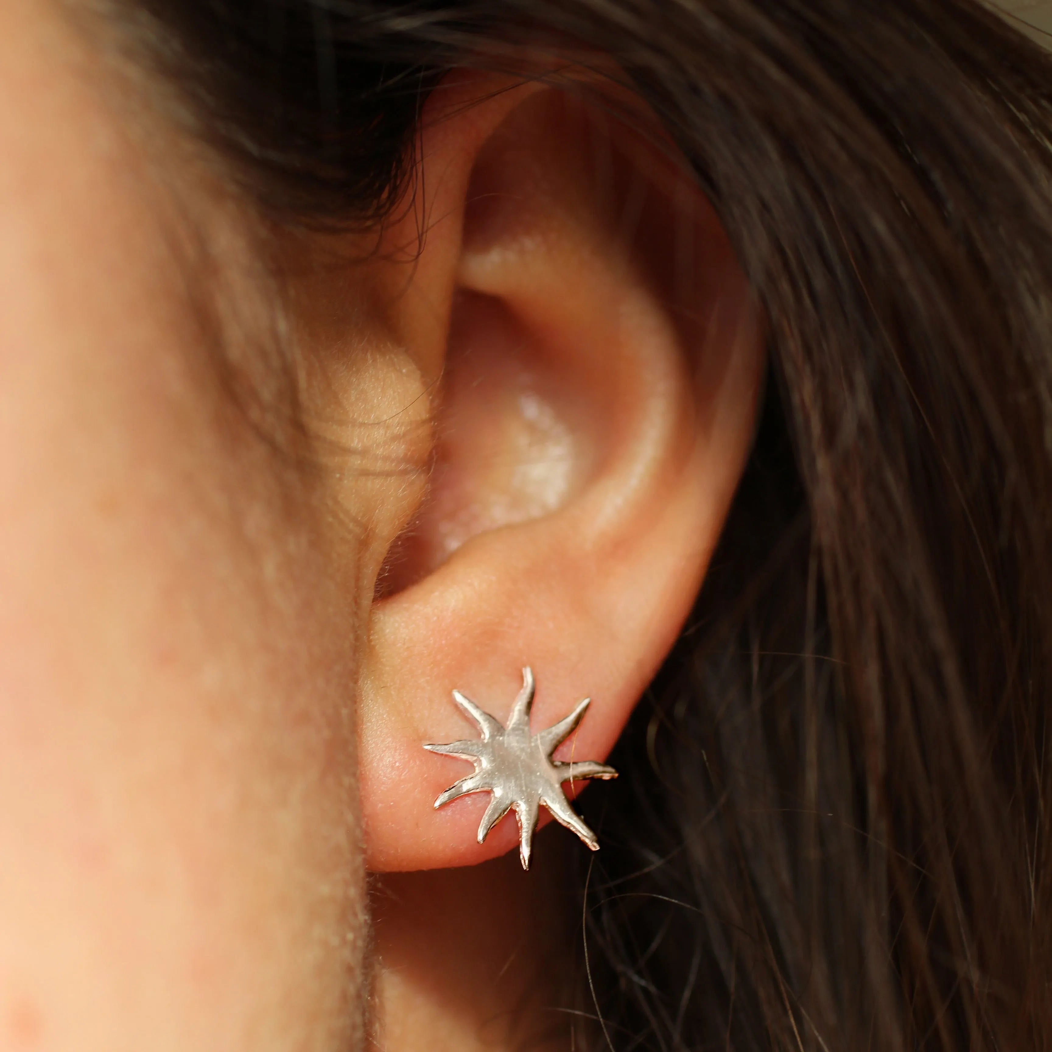 sun shaped stud earring in silver worn in ear with brunette hair in background
