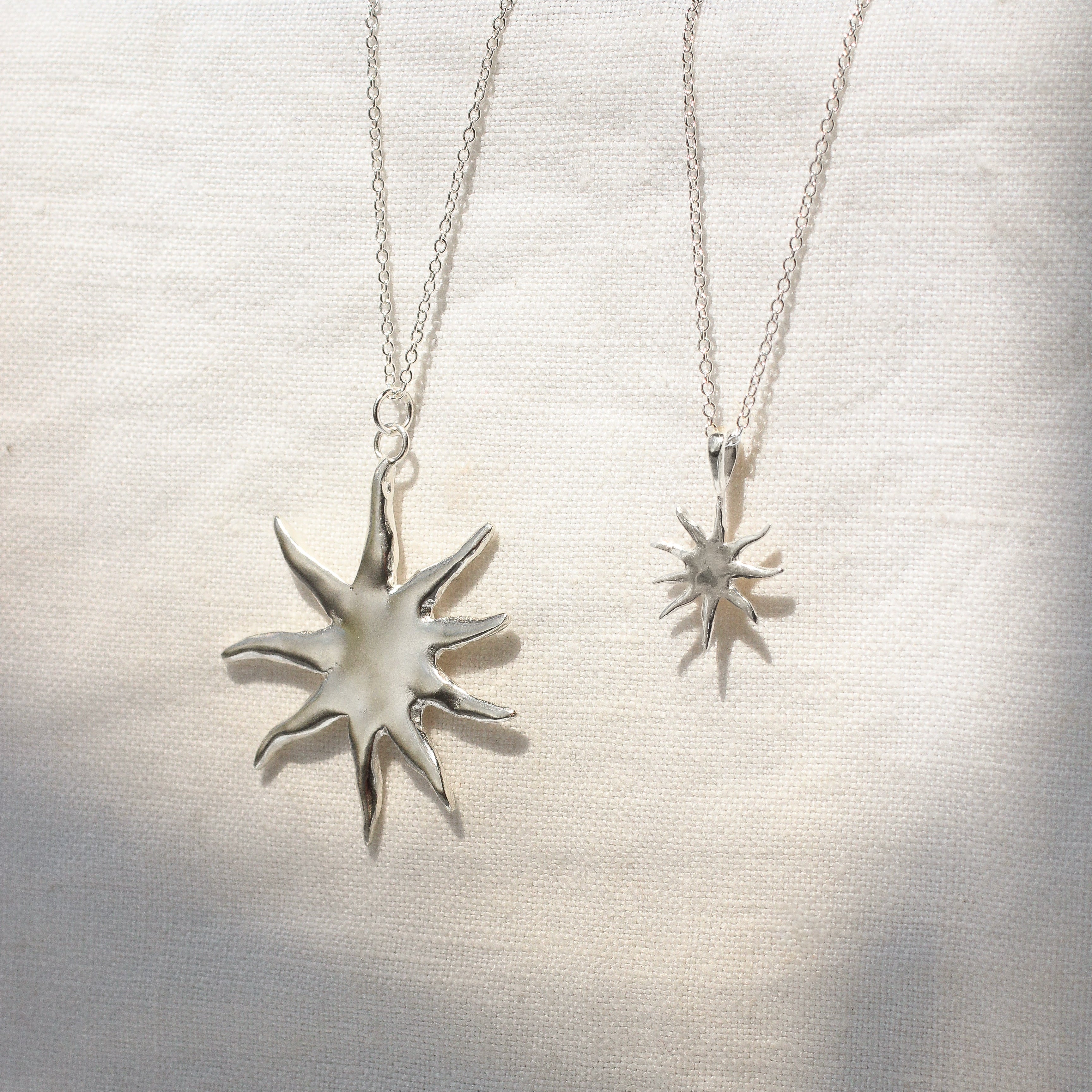 Two silver sun-shaped pendants on delicate chains against a textured white background
