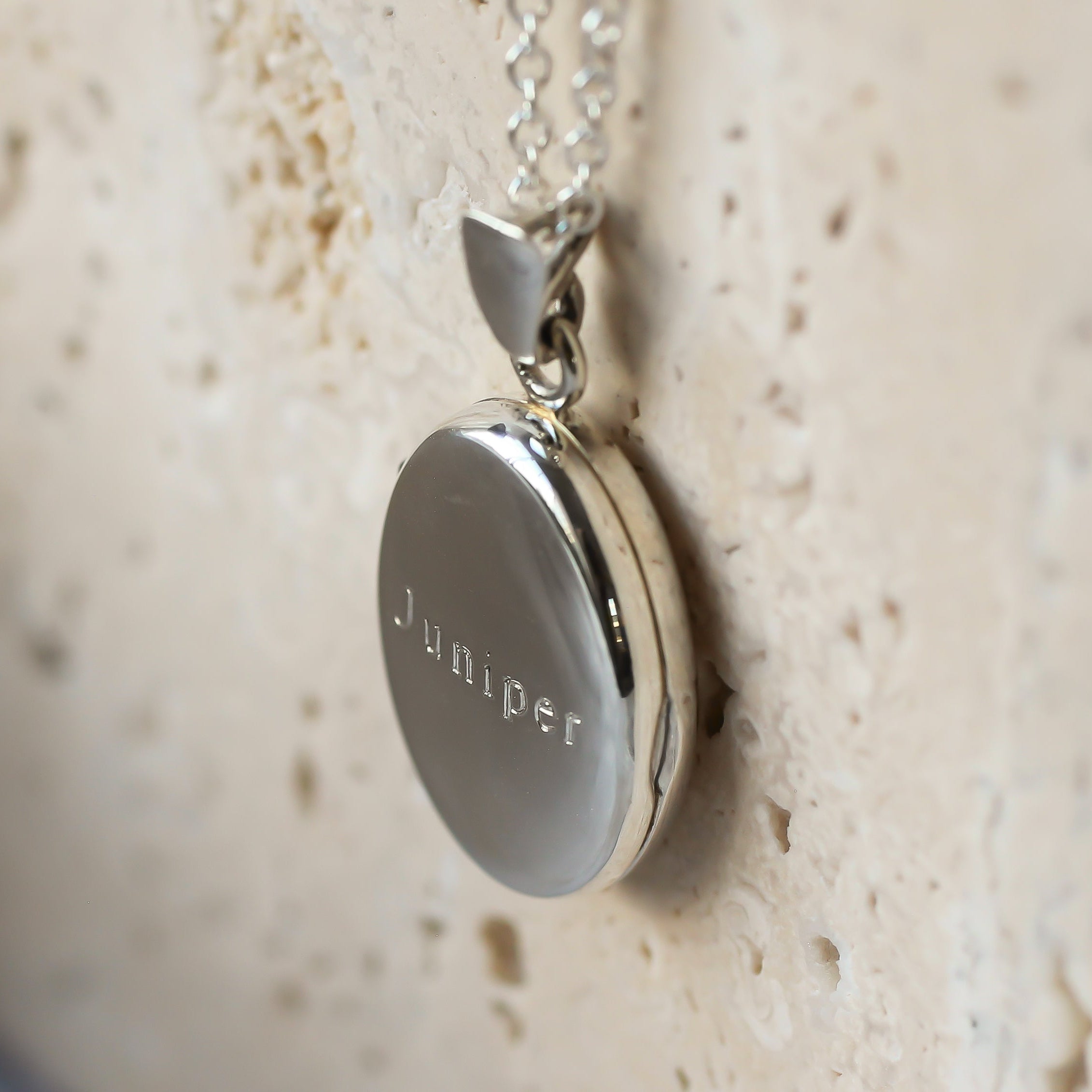 A silver round locket pendant engraved with the name 'Juniper' on a silver chain, resting against a textured beige stone background.