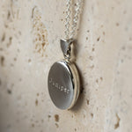 A silver round locket pendant engraved with the name 'Juniper' on a silver chain, resting on a textured beige stone surface.
