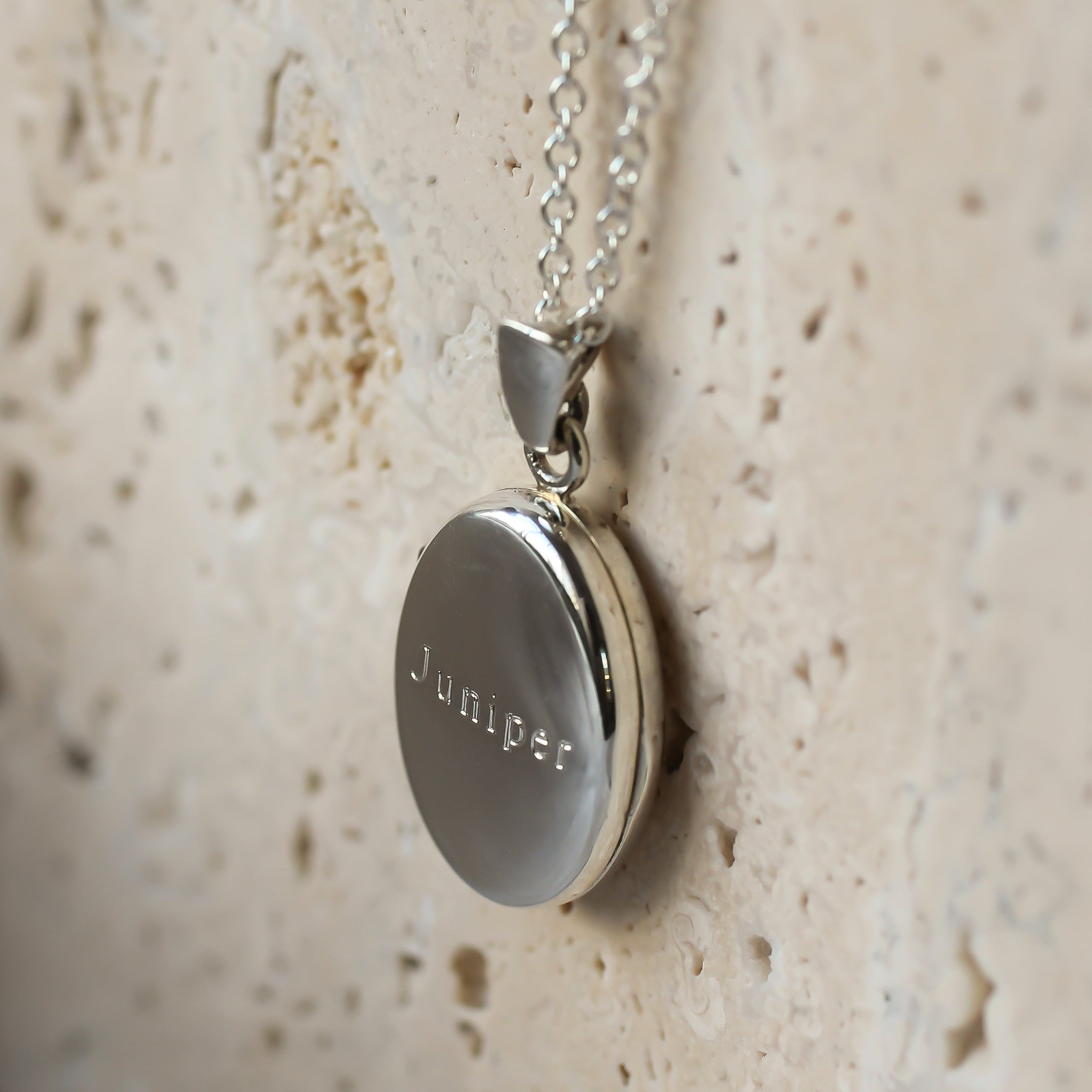 A silver round locket pendant engraved with the name 'Juniper' on a silver chain, resting on a textured beige stone surface.