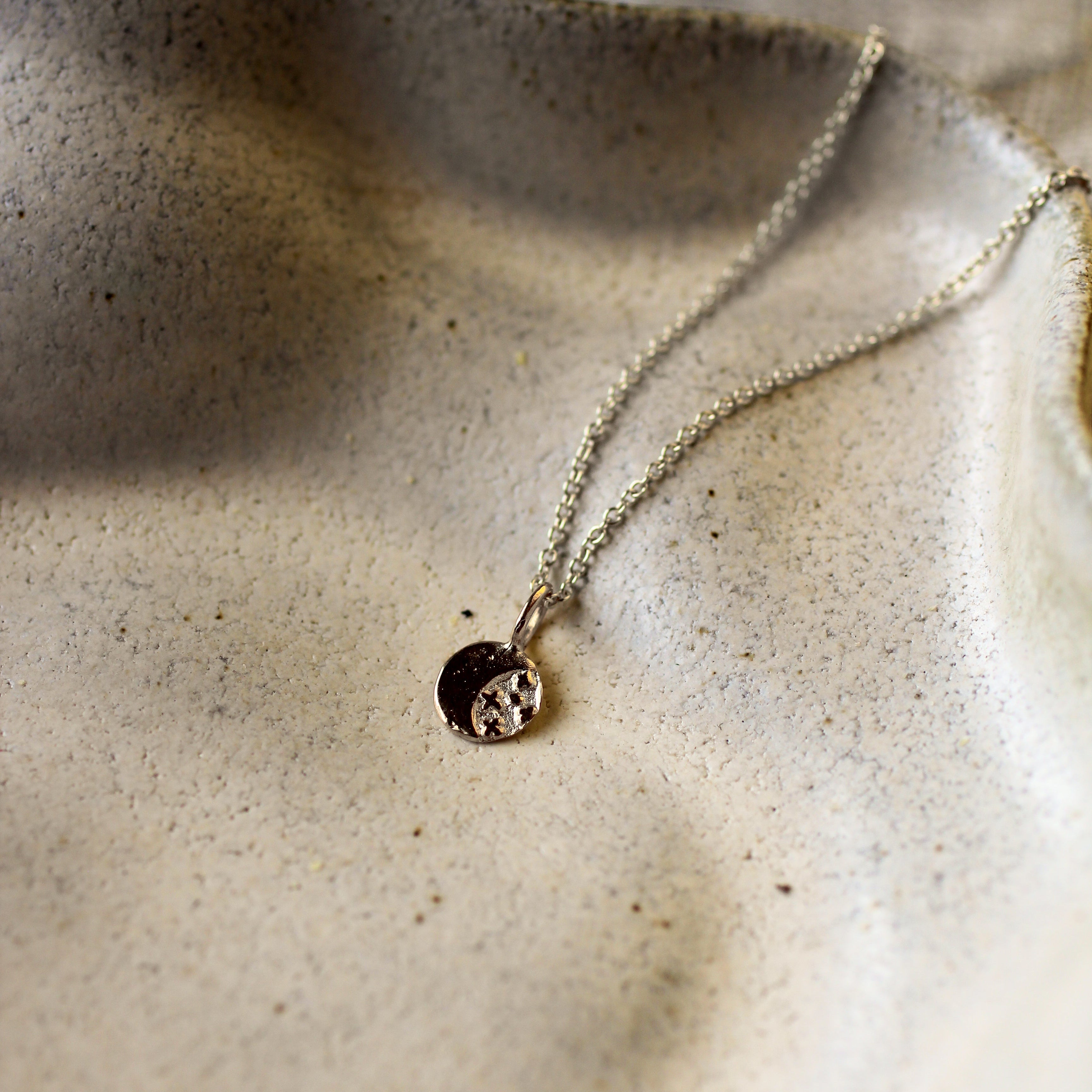 A delicate silver chain necklace with a small circular pendant resting on a textured beige ceramic dish.