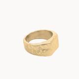 A textured, chunky signet-style ring in a warm gold finish, photographed on a clean, solid off-white background.