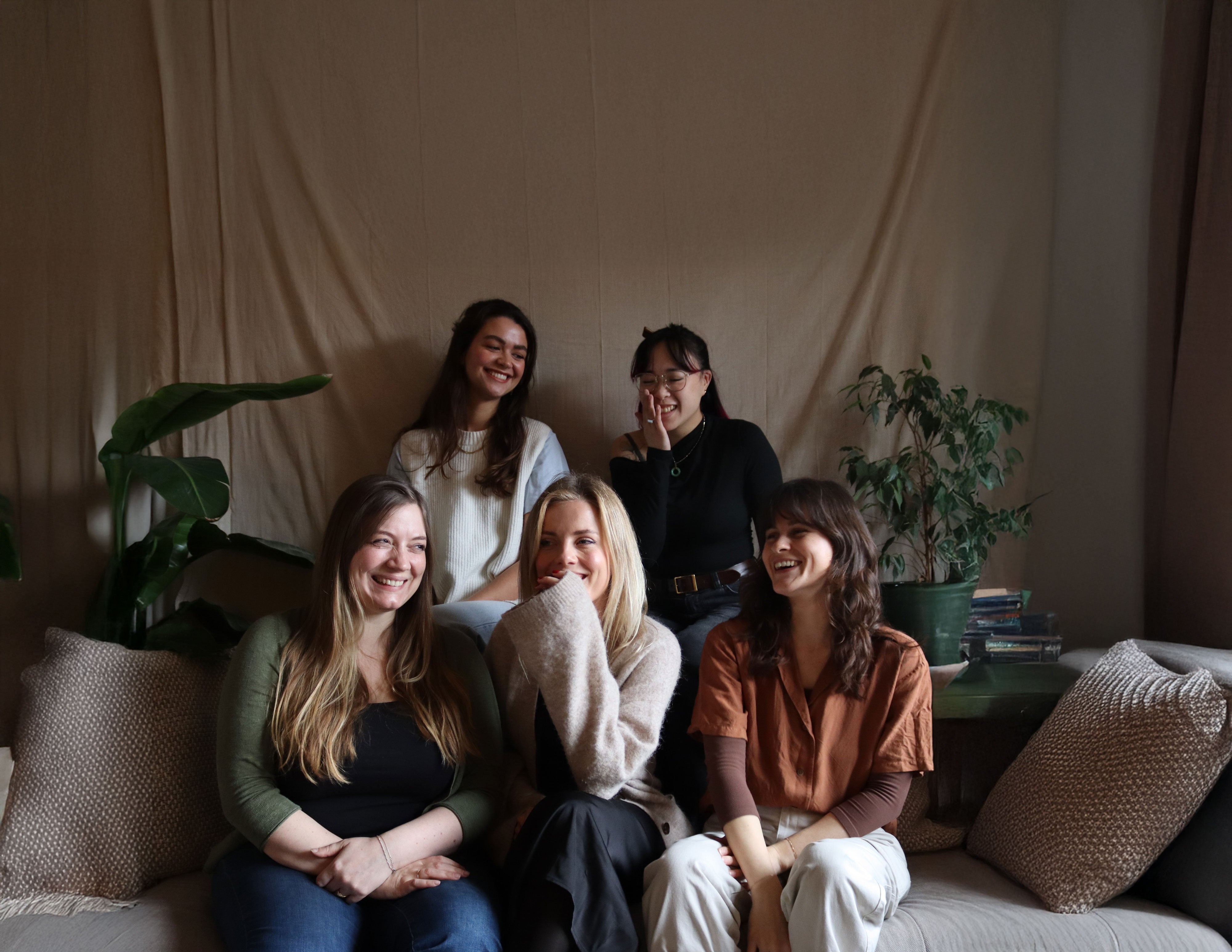 Five women sitting on a couch in a cozy room with plants and a neutral background