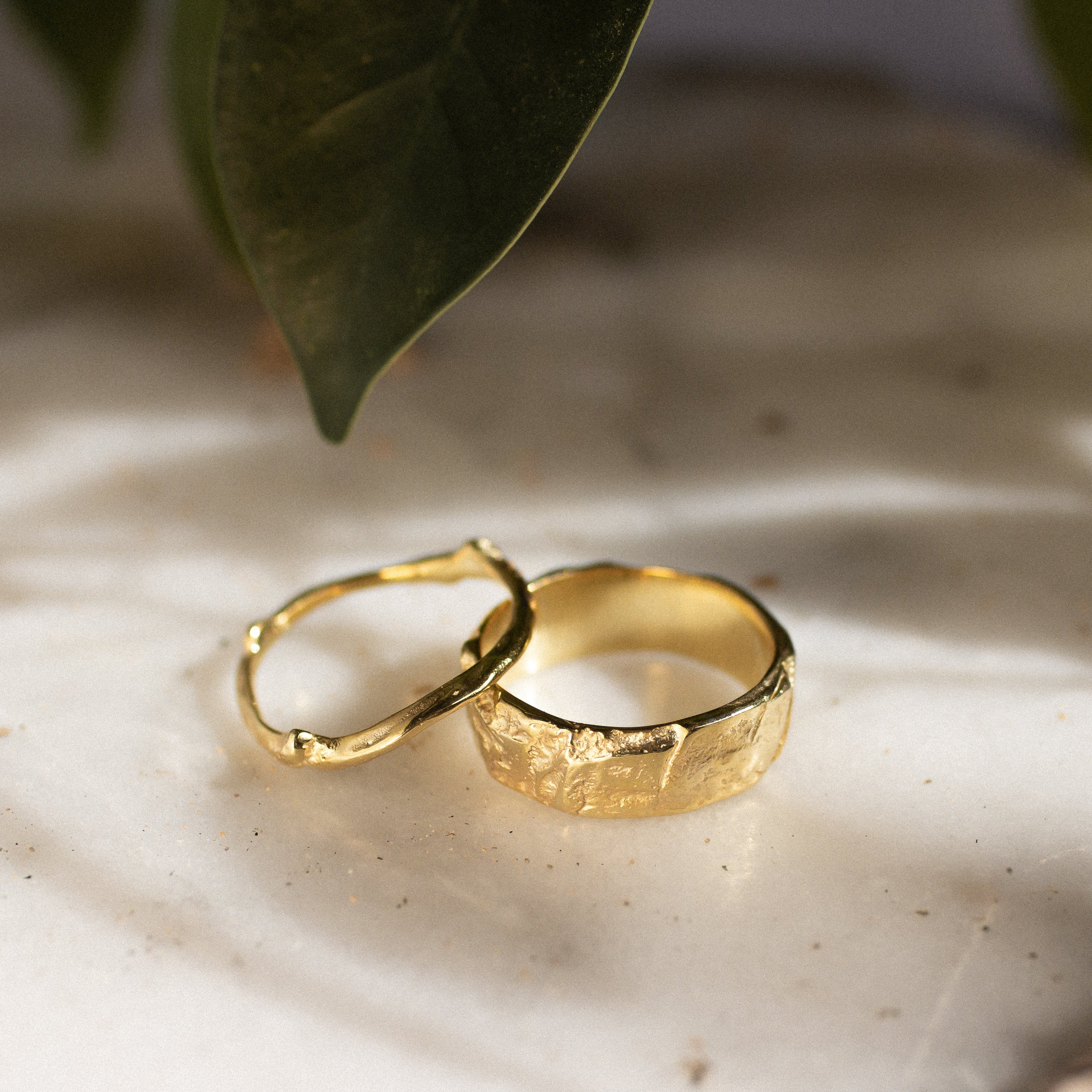 a thick tree bark wedding ring and slim tree branch wedding ring on a textured surface with shadows and leaves in the background