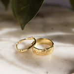 a thick tree bark wedding ring and slim tree branch wedding ring on a textured surface with shadows and leaves in the background