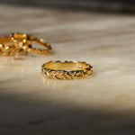 A gold ring with a textured, braided or rope-like outer band, resting on a light marble surface under warm, dramatic lighting.