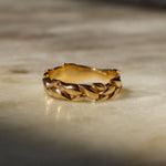 A gold ring with a textured, leaf-like braided pattern, resting on a light beige marble surface with soft shadows.