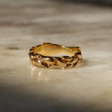 A gold ring with a textured, leaf-like braided pattern, resting on a light beige marble surface with soft shadows.