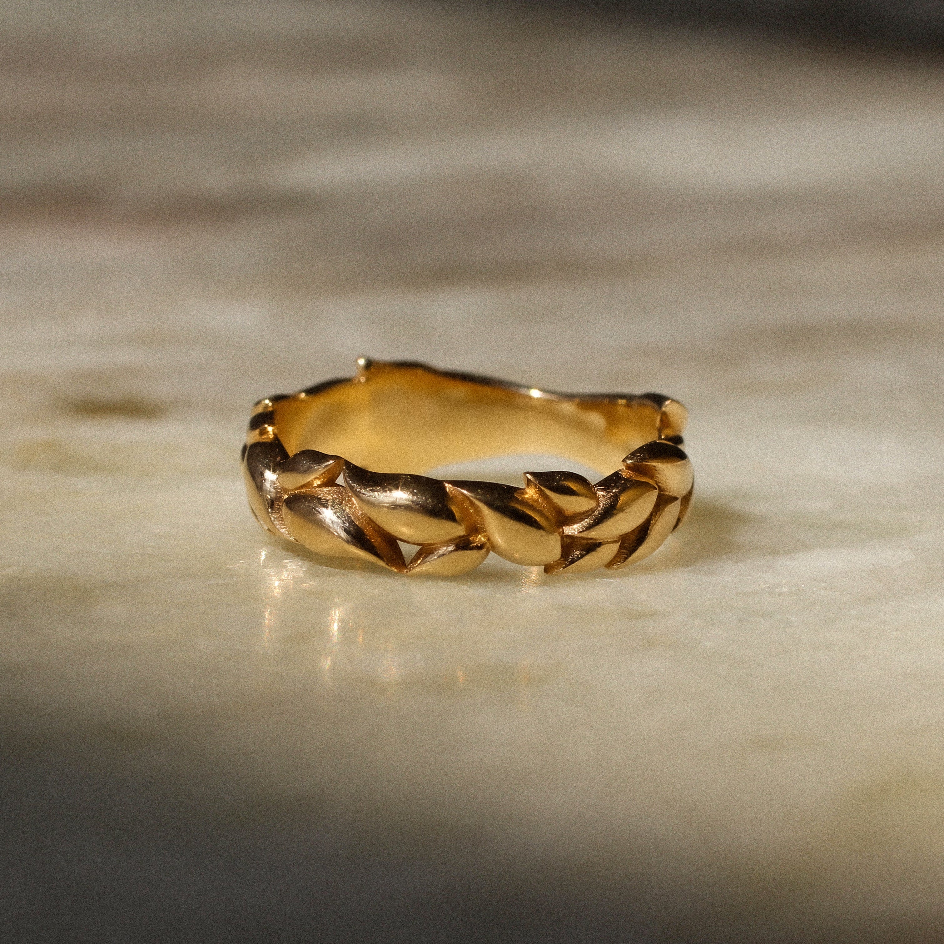 A gold ring with a textured, leaf-like braided pattern, resting on a light beige marble surface with soft shadows.
