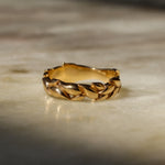 A gold ring with a textured, leaf-like braided pattern, resting on a light beige marble surface with soft shadows.