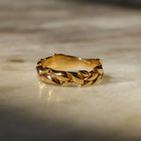 A gold ring with a textured, leaf-like braided pattern, resting on a light beige marble surface with soft shadows.