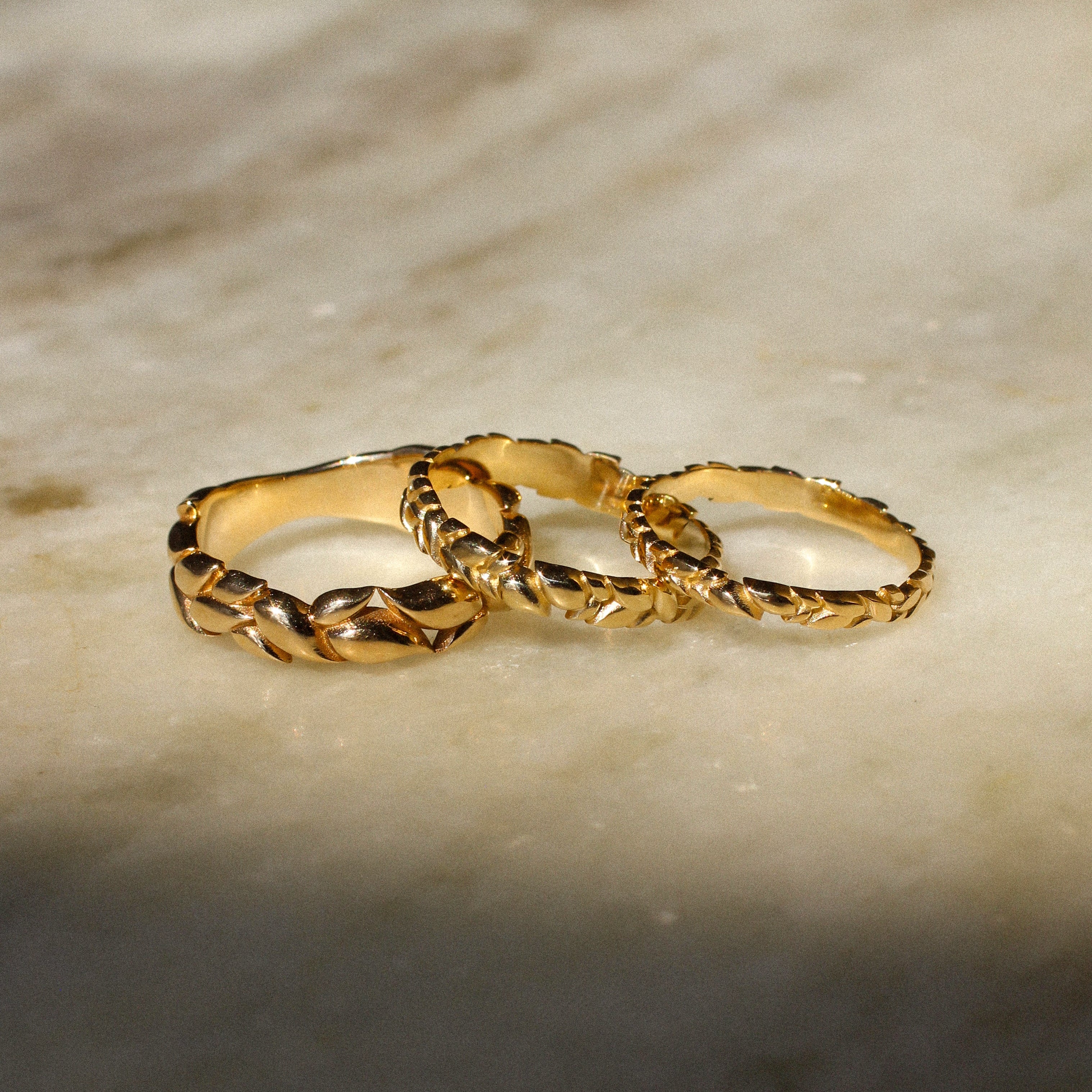 Three interlocking textured gold rings displayed on a light beige marble surface with soft shadows.