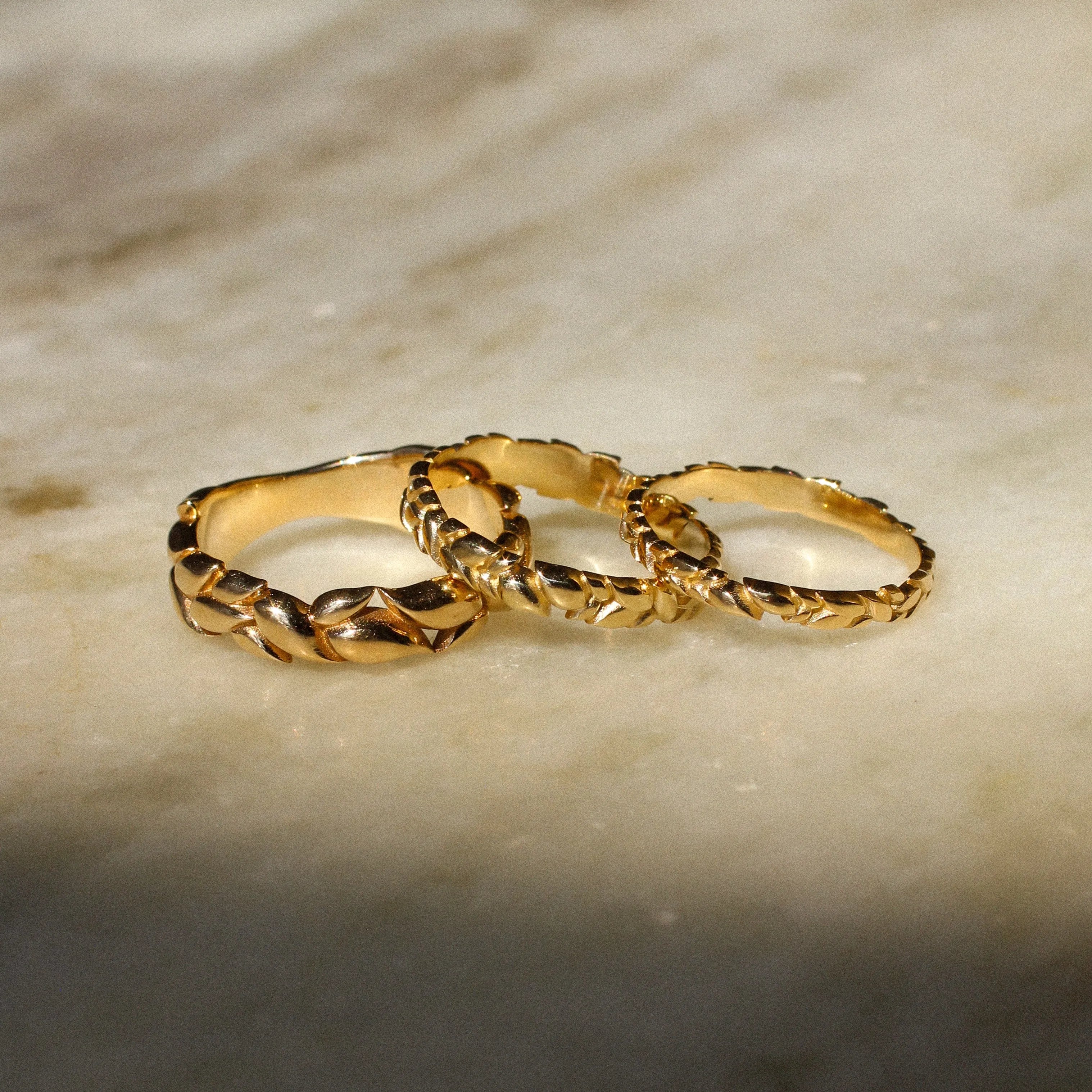Three interlocking textured gold rings displayed on a light beige marble surface with soft shadows.