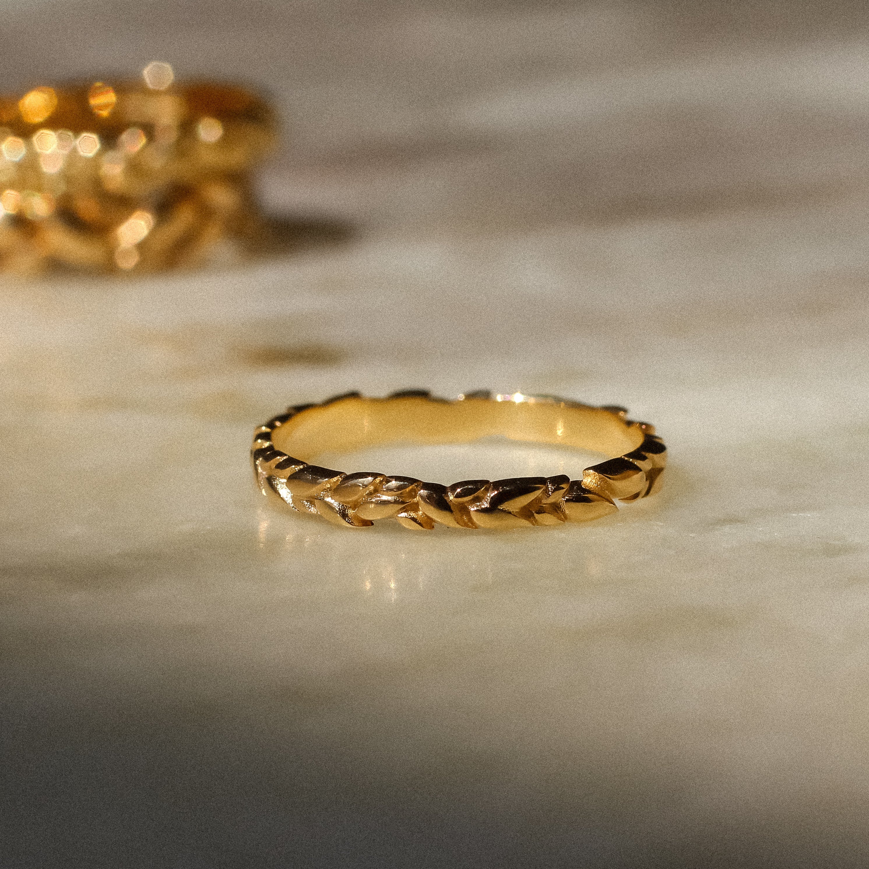 A gold ring with a textured, braided or rope-like band design, resting on a light marble surface with a second matching ring blurred in the background.