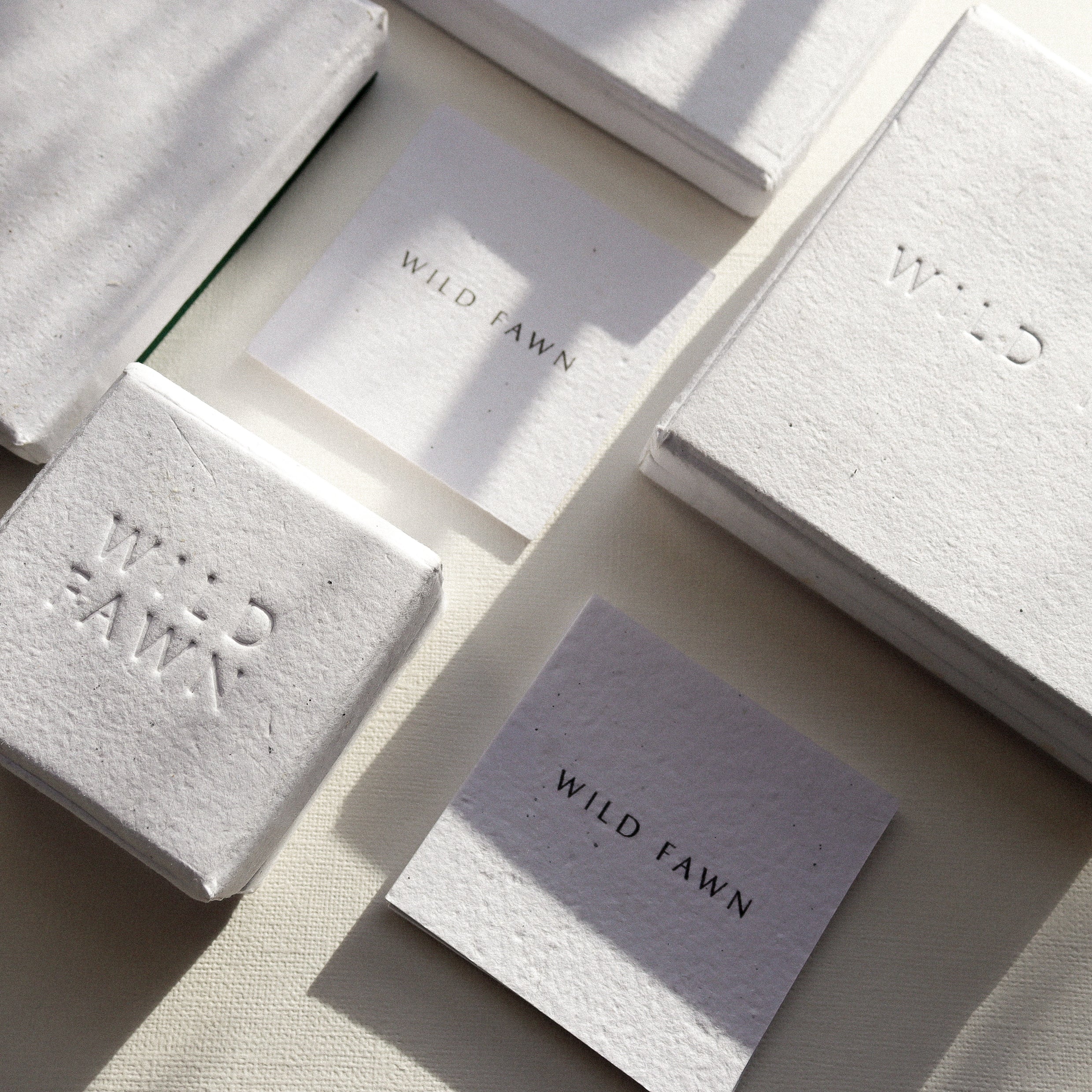 A styled flat-lay of textured white paper cards and matching paper blocks embossed with the brand name 'WILD FAWN', arranged on a neutral beige surface with soft diagonal shadows.